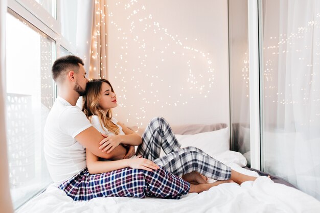 Impresionante chica en pijama perezoso posando con novio. Adorable joven tumbado en la cama junto a su marido en fin de semana.