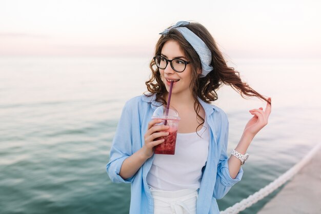 Impresionante chica rizada sosteniendo jugo de cereza y jugando con su cabello oscuro durante el paseo por el muelle