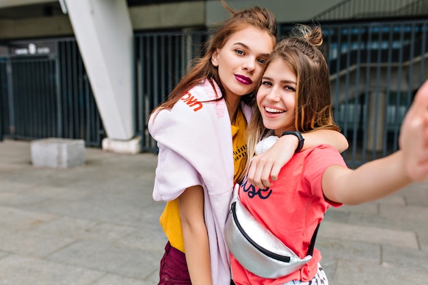 Impresionante chica rubia inspirada con bolso plateado haciendo selfie con hermana antes de ir de compras en verano