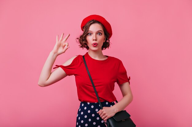 Impresionante chica sorprendida en camiseta roja posando divertida. Filmación en interiores de espectacular modelo femenino en boina expresando emociones de sorpresa.