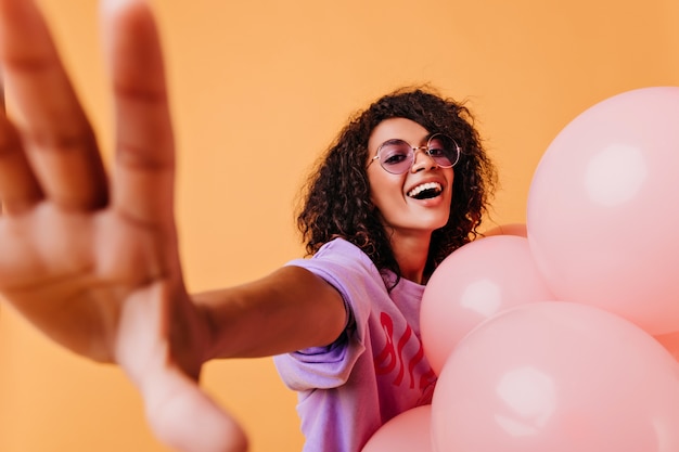 Impresionante cumpleañera en gafas de moda haciendo selfie. mujer africana con lindos globos rosados riendo en amarillo.