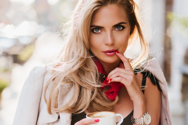 Impresionante dama bronceada posando juguetonamente tocando los labios con el dedo. Chica bonita rubia sosteniendo una taza de té y mirando con interés