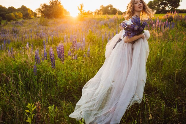 Impresionante dama en vestido blanco se encuentra con ramo en el campo