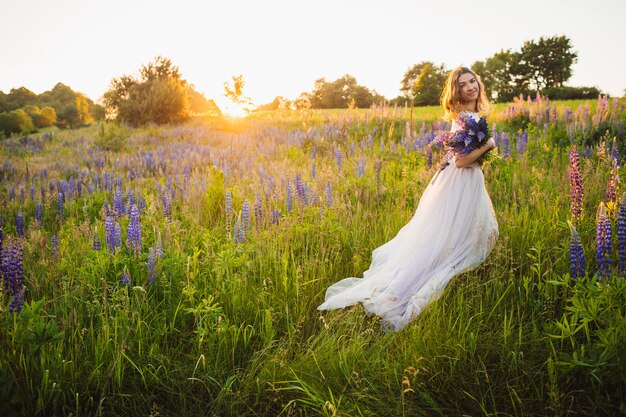 Impresionante dama en vestido blanco se encuentra con ramo en el campo