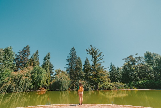 Impresionante foto de una mujer de pie frente a un río en medio de un bosque en un día caluroso