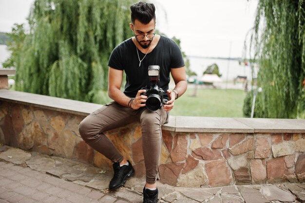 Impresionante y hermoso fotógrafo de hombre macho de barba árabe alto con gafas y camiseta negra con cámara profesional en las manos