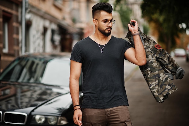 Impresionante hermoso hombre macho alto de barba árabe con gafas y camiseta negra caminando contra un coche de negocios