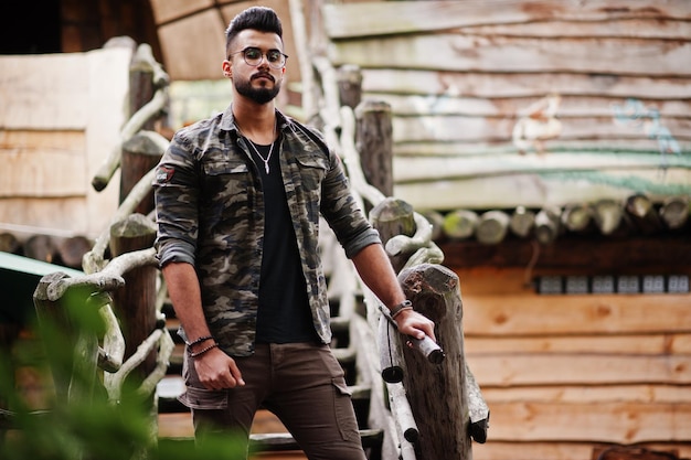 Foto gratuita impresionante hermoso hombre macho alto de barba árabe con gafas y chaqueta militar posado al aire libre contra escaleras de madera