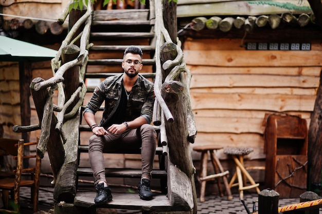 Impresionante hermoso hombre macho alto de barba árabe con gafas y chaqueta militar posado al aire libre contra escaleras de madera