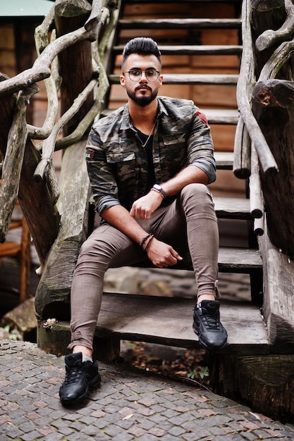 Impresionante hermoso hombre macho alto de barba árabe con gafas y chaqueta militar posado al aire libre contra escaleras de madera