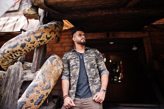 Foto gratuita impresionante hermoso hombre macho alto de barba árabe con gafas y chaqueta militar posado al aire libre contra pitón serpiente