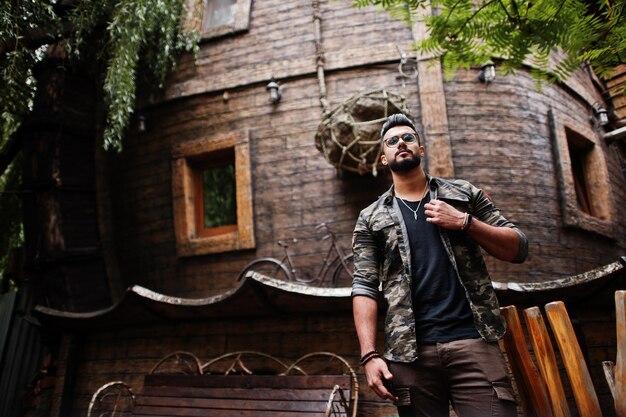 Impresionante hombre macho macho de barba árabe alto con gafas y chaqueta militar posado al aire libre contra la casa de madera