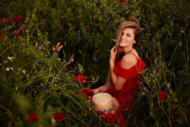 Impresionante joven en vestido rojo se sienta en el campo verde con amapolas rojas