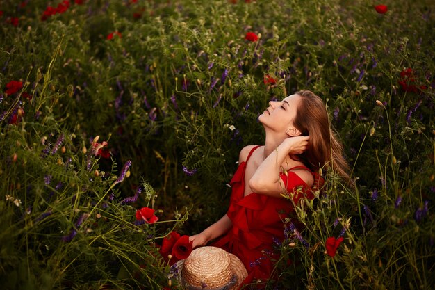 Impresionante joven en vestido rojo se sienta en el campo verde con amapolas rojas