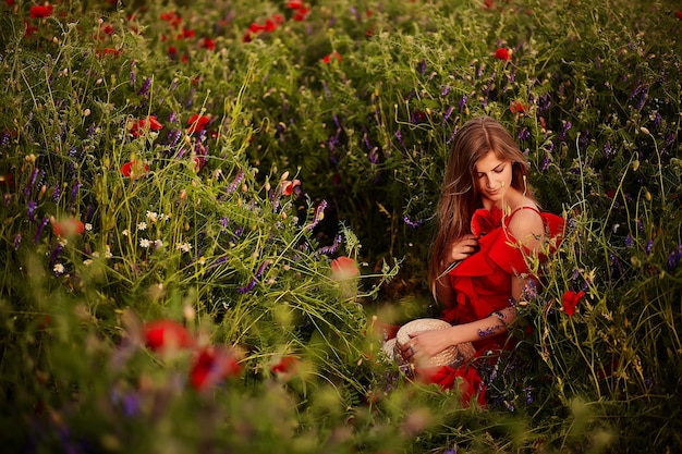 Impresionante joven en vestido rojo se sienta en el campo verde con amapolas rojas