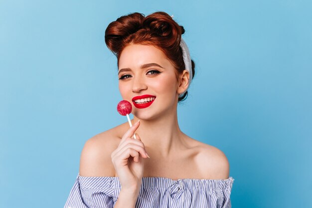 Impresionante jovencita caucásica lamiendo piruleta. Chica pinup feliz comiendo caramelos en el espacio azul.