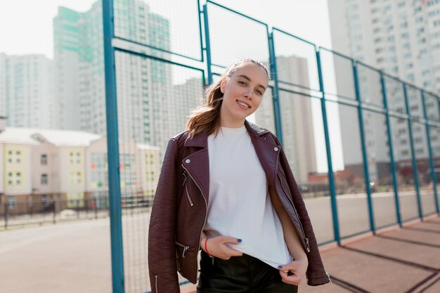 impresionante linda chica vestida con camiseta blanca y chaqueta burdeos está posando en la cámara con emociones felices al aire libre en el fondo del área recreativa de la ciudad