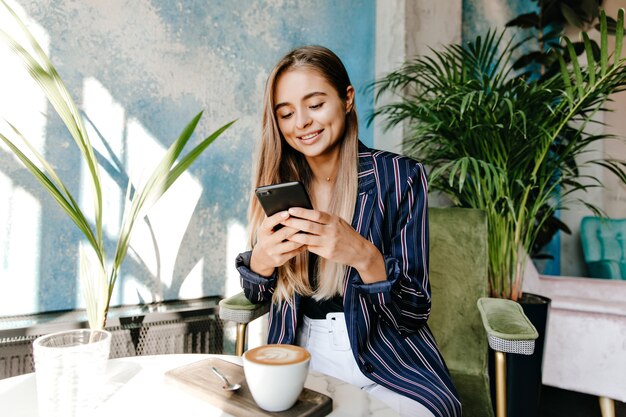 Impresionante mensaje de texto de mujer blanca mientras bebe capuchino. Adorable chica europea en chaqueta descansando en el café.