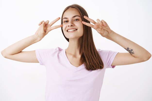 Impresionante mujer caucásica feliz y despreocupada con tatuaje en el brazo que muestra signos de discoteca o paz cerca de los ojos y sonriendo con mirada alegre bailando pasando los fines de semana divertidos y entretenidos