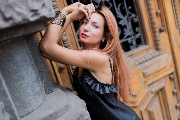 Impresionante mujer elegante rubia en vestido de noche negro posando en la ciudad. Labios rojos, pelos rojos, accesorios inusuales.