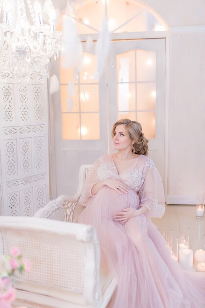 Impresionante mujer embarazada en vestido rosa descansa en el sofá en una habitación blanca