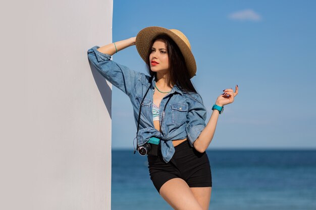 Impresionante mujer con figura en pantalones cortos y sombrero de paja posando en la playa.