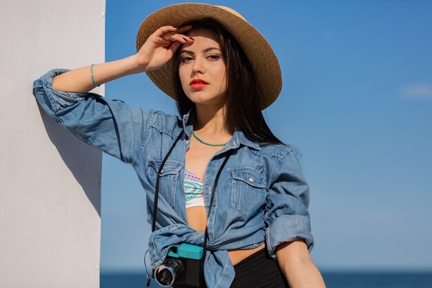 Impresionante mujer con figura en pantalones cortos y sombrero de paja posando en la playa.