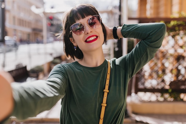 Impresionante mujer joven con una hermosa sonrisa disfrutando del día de primavera Toma al aire libre de una chica morena refinada con atuendo moderno caminando por la calle y riendo