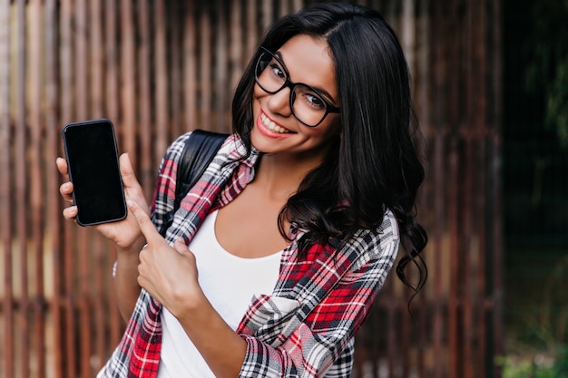 Impresionante mujer latina rizada mostrando nuevo teléfono. Retrato al aire libre de la adorable chica morena con gafas posando en la pared de madera con smartphone.