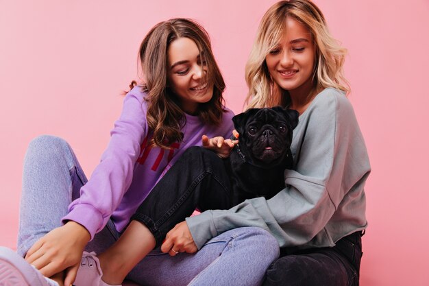 Impresionante mujer sosteniendo cachorro negro con sonrisa encantadora. Filmación en interiores de adorables hermanas que expresan felicidad durante la sesión de retratos con un pequeño bulldog.