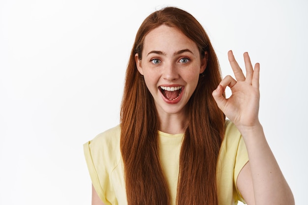 Impresionante, muy bueno. La chica pelirroja sonriente muestra un gesto correcto, sin problemas, elogios y elogios, dice sí, aprueba y le gusta algo, fondo blanco.