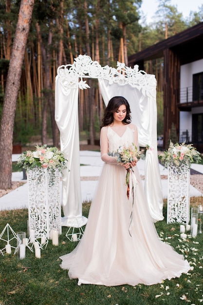 Impresionante novia con un vestido beige se encuentra ante un altar de bodas fuera