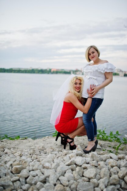 Impresionante novia en vestido rojo posando con su dama de honor embarazada en la orilla del lago