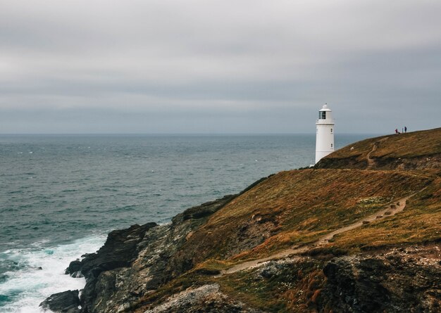 Impresionante vista de un faro en una colina cubierta de hierba junto al océano en un día nublado