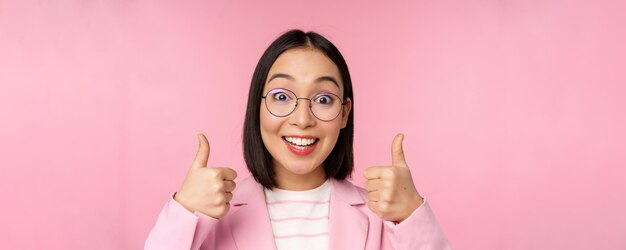 Impresionantes felicitaciones Cara de emocionada mujer de negocios asiática con gafas sonriendo complacida mostrando los pulgares hacia arriba en señal de aprobación de pie sobre fondo rosa