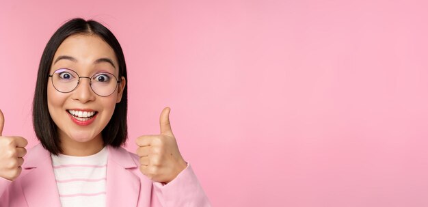 Impresionantes felicitaciones Cara de emocionada mujer de negocios asiática con gafas sonriendo complacida mostrando los pulgares hacia arriba en señal de aprobación de pie sobre fondo rosa