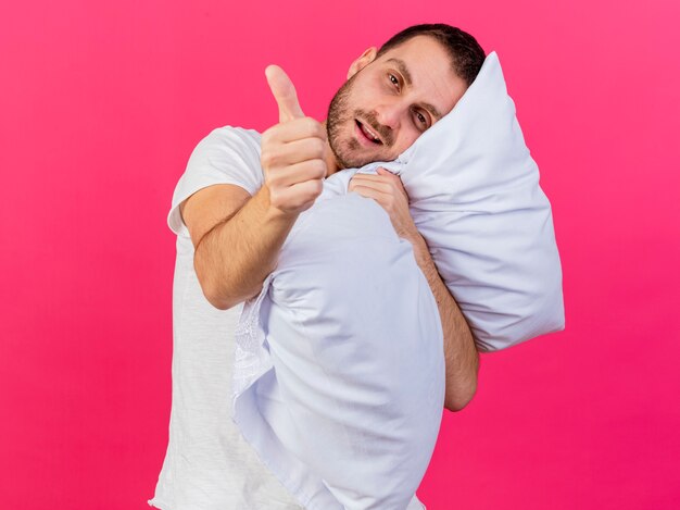 Foto gratuita inclinando la cabeza joven enfermo abrazó la almohada y mostrando el pulgar hacia arriba aislado en rosa