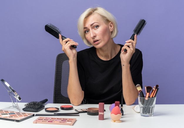 Inclinar la cabeza hermosa joven se sienta a la mesa con herramientas de maquillaje sosteniendo peines aislados en la pared azul