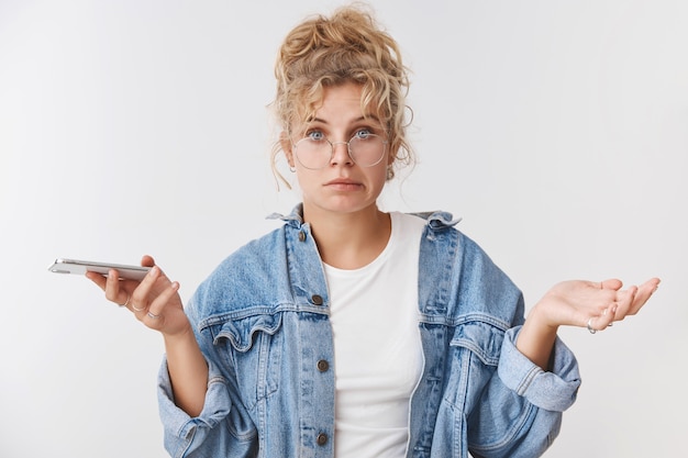 Incómodo inseguro desorientado lindo torpe asistente femenino rubio europeo usar anteojos moño desordenado sonriendo inseguro extendió las manos hacia los lados sin darse cuenta sosteniendo el teléfono inteligente no estoy seguro de cómo resolver el problema