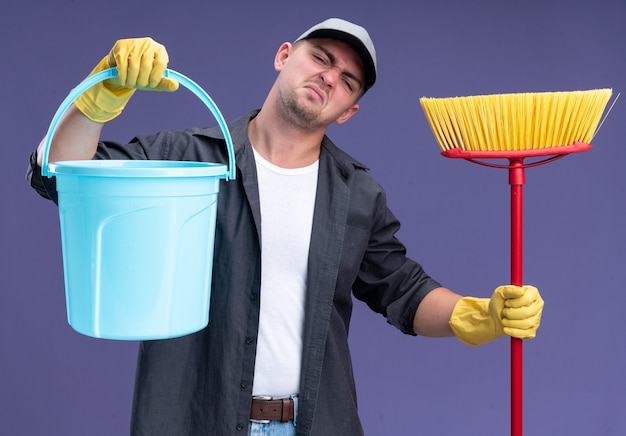 Foto gratuita incompatible inclinando la cabeza joven guapo chico de limpieza con camiseta y gorra con glaves sosteniendo un cubo con un trapeador aislado en la pared púrpura