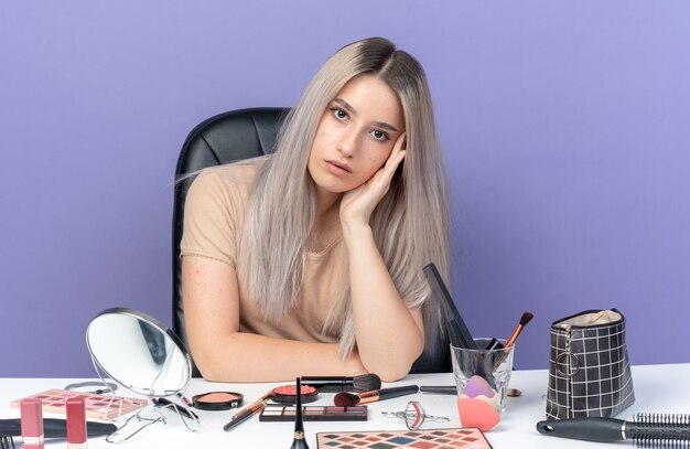 Inconforme la cabeza inclinada joven hermosa niña se sienta a la mesa con herramientas de maquillaje poniendo la mano en la mejilla aislada sobre fondo azul