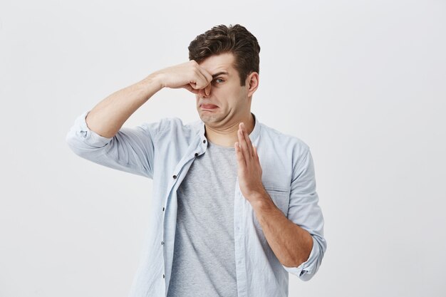 Inconformista masculino caucásico con cabello oscuro vestido con una camisa azul claro sobre una camiseta gris que se pellizca la nariz debido al mal olor de algo sucio y apestoso, con expresión de disgusto.