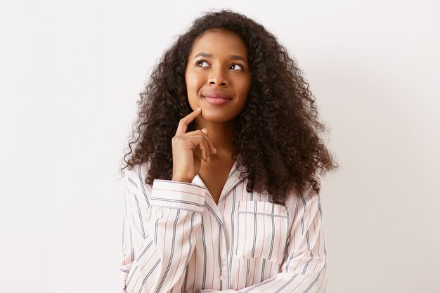 Increíble adorable niña de origen afroamericano con expresión facial pensativa de ensueño, mirando hacia arriba y sonriendo, sosteniendo el dedo índice en su barbilla, pensando en planes de vacaciones y futuro emocionante