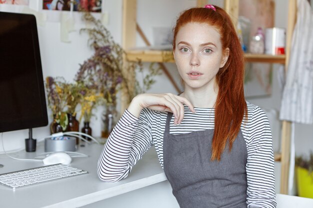 Increíble artista femenina profesional con pecas y cabello largo y pelirrojo sentada en la computadora en una mesa blanca en su moderno taller, con una mirada pensativa, sumida en pensamientos, absorta en ideas creativas