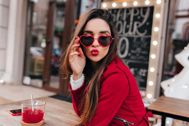 Increíble chica blanca en gafas de corazón de moda posando con expresión de cara de besos