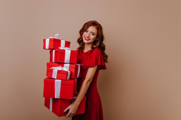 Increíble chica con cabello ondulado de jengibre sosteniendo regalos rojos Foto de estudio de modelo femenino emocional posando con regalos de navidad