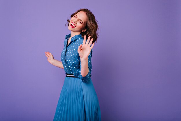 Increíble chica caucásica en vestido azul vintage sonriendo con los ojos cerrados Elegante jovencita con peinado ondulado corto bailando en la pared púrpura.