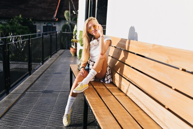 Increíble chica delgada en zapatos amarillos posando en banco de madera. Tiro al aire libre de mujer rubia bronceada en ropa casual, pasar tiempo en la ciudad.