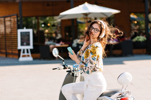 Increíble chica elegante con teléfono en scooter esperando novio que fue a tomar un café en la cafetería