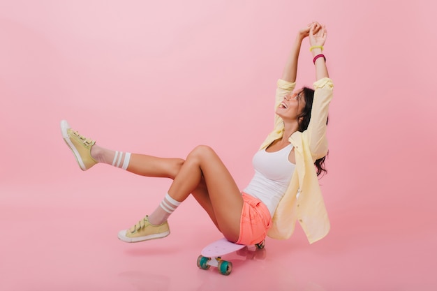 Increíble chica latina en calcetines de rayas rosas disfrutando de la vida y sonriendo. Agraciada mujer joven con cabello oscuro posando con las manos arriba, sentada en patineta